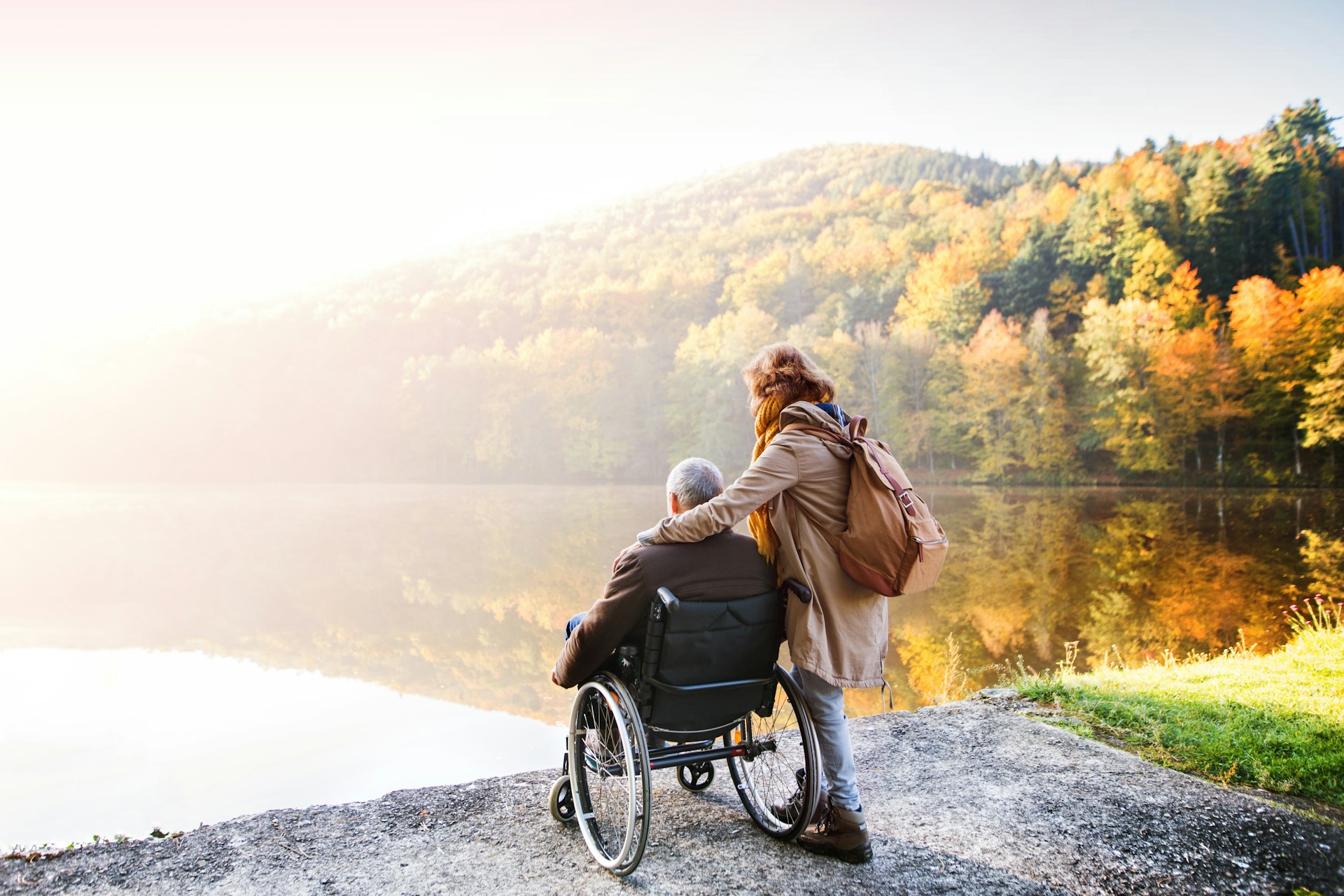 Pflegebedürftiger alter Mann im Rollstuhl schaut mit seiner jungen Tochter auf einen See bei Sonnenuntergang im Herbst.
